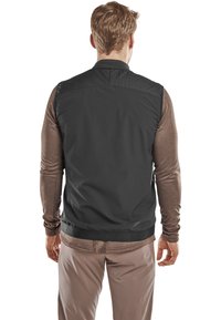 Zwarte mouwloze vest met een soepele textuur, hoge kraag en zachte voering, gedragen over een bruin langemouwshirt en beige broek.
