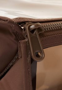 Borsa di stoffa marrone con cerniera e un tirante arrotondato, cuciture visibili lungo i bordi, che mette in mostra una superficie liscia e riflettente sottostante.