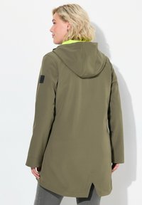 Cappotto impermeabile verde oliva con cappuccio. Presenta una patch nera, tasche laterali e un orlo a incastro. Tessuto liscio e testurizzato.