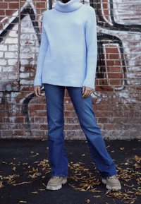 Person som bär en ljusblå polo-tröja, blå jeans och beiga klumpiga skor står på en asfalt täckt av löv nära en tegelvägg med graffiti.