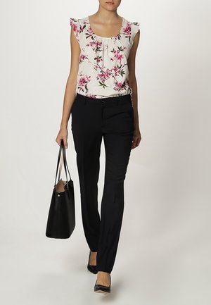 Witte bloemenblouse met volants aan de mouwen, gecombineerd met zwarte, op maat gemaakte broek. Een zwarte tote bag en flats met puntige neuzen maken de look compleet.