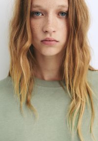 Lichtgroene katoenen t-shirt met een ronde halslijn, gedragen met los, golvend blond haar dat het gezicht omlijst; blauwe ogen en lichte sproeten zijn zichtbaar.