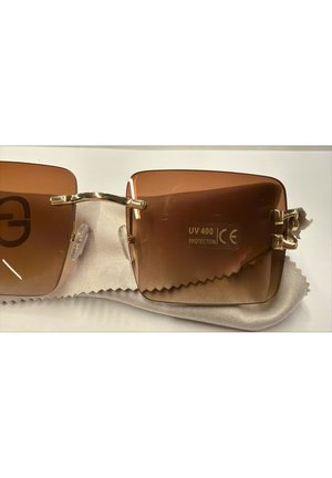 Lunettes de soleil carrées marron avec monture dorée, plaquettes de nez, et étiquette "Protection UV 400" sur le verre droit reposant sur un tissu clair.