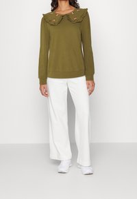 Felpa verde oliva con ampio colletto arricciato, ricamo floreale, polsini e orlo a coste; abbinata a pantaloni bianchi a gamba larga e sneakers bianche.