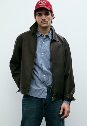 Fiatal férfi piros sapkában, fekete dzsekiben, kék csíkos ing alatt és kék farmerben, sima világos háttér előtt áll.