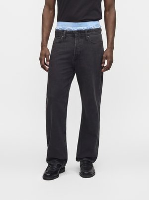 Homme portant un jean noir décontracté, des chaussures noires et une chemise noire, avec un sous-vêtement bleu clair visible au-dessus de la ceinture.