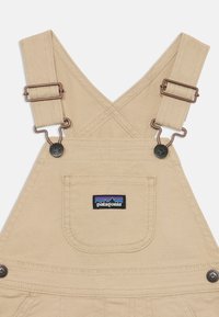 Salopette en toile beige avec des bretelles ajustables, des accessoires en bronze, une poche avant et un patch logo Patagonia. Présente des coutures renforcées.