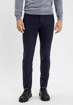 Man draagt zwarte slim-fit broek, zwarte sneakers met veters en een lichtgrijze trui met lange mouwen, staand op een witte achtergrond.