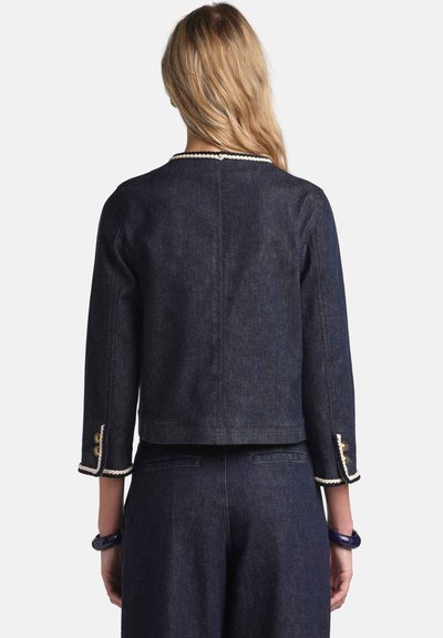 Mørk denimjakke med trekvartlange ærmer, udsmykket med dekorative gyldne knapper og hvide kanter langs manchetterne og halsudskæringen.