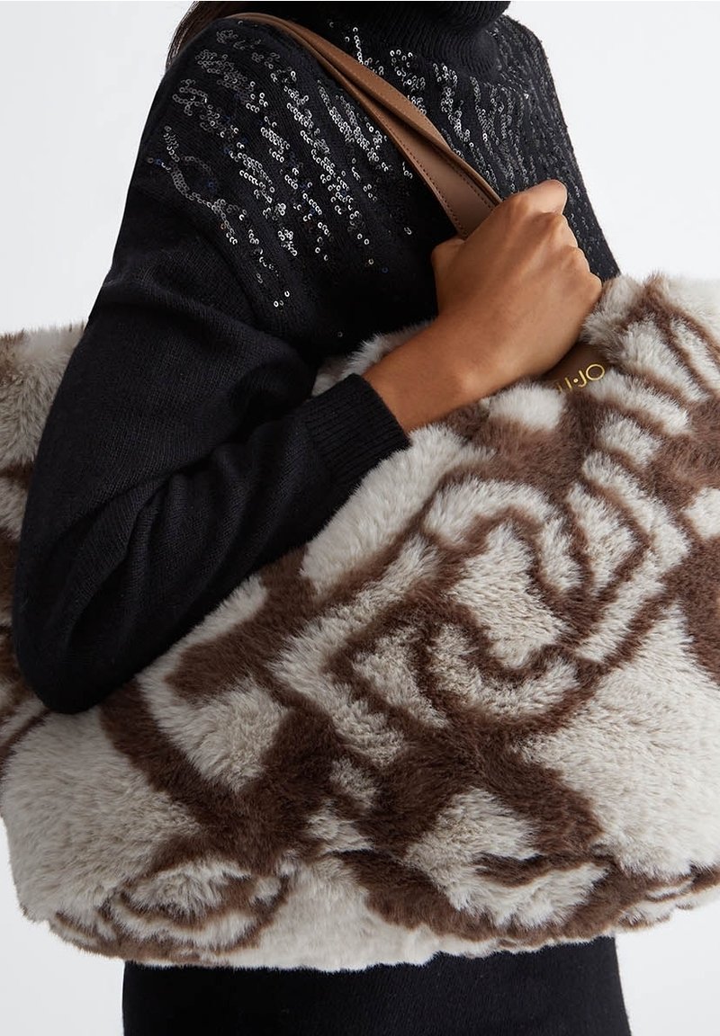 Personne portant un pull noir à sequins porte un grand sac en fausse fourrure beige et marron à motifs avec des bretelles en cuir marron sur l'épaule.