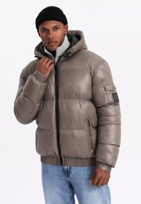 Giubbotto imbottito grigio con zip frontale, polsini a costine e cappuccio. Presenta una tasca sulla manica con un patch nero. Indossato sopra un top di colore chiaro.
