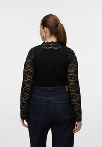 Haut en dentelle noire à manches longues, décolleté à bord festonné, associé à un jean en denim sombre avec poches arrière et détails de couture.