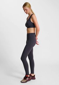 Czarny sportowy biustonosz na cienkich ramiączkach, w połączeniu z czarnymi legginsami z wysokim stanem. Modelka w czerwono-szarych butach sportowych, widok z profilu.