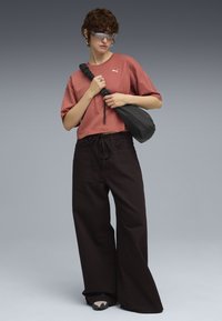Lyserød kortærmet t-shirt med Puma-logo, sorte bredde benbukser med snøre, og en sort sling taske; sølvtonede solbriller, minimale accessories.