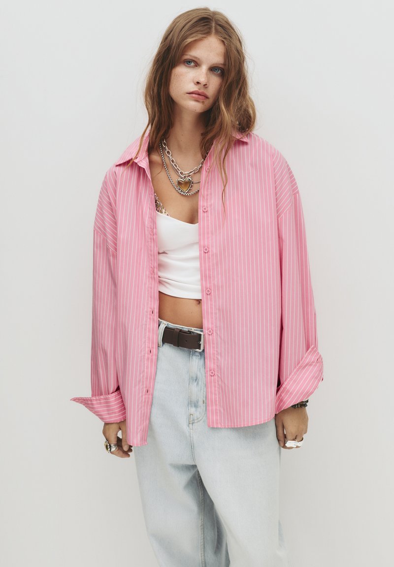 Camicione oversize a righe rosa con chiusura a bottoni, realizzato in tessuto leggero, abbinato a jeans azzurri chiari e collane in argento sovrapposte.