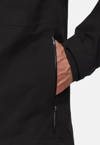 Veste imperméable noire avec une poche latérale zippée, un tissu texturé et des poignets de manches ajustables. Présente un design minimaliste et des lignes épurées.