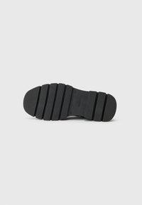Schwarze Schuhsohle mit strukturiertem Grip und horizontalen Rillen, fotografiert vor einem schlichten hellgrauen Hintergrund.