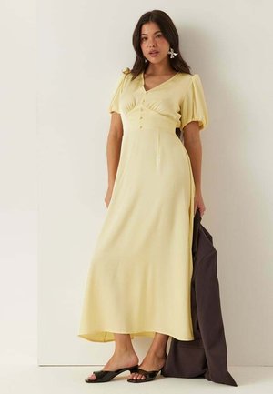 Vrouw draagt een lichtgele midi-jurk met pofmouwen en knopen, houdt een donkere jas vast en draagt zwarte open-toe hakken.