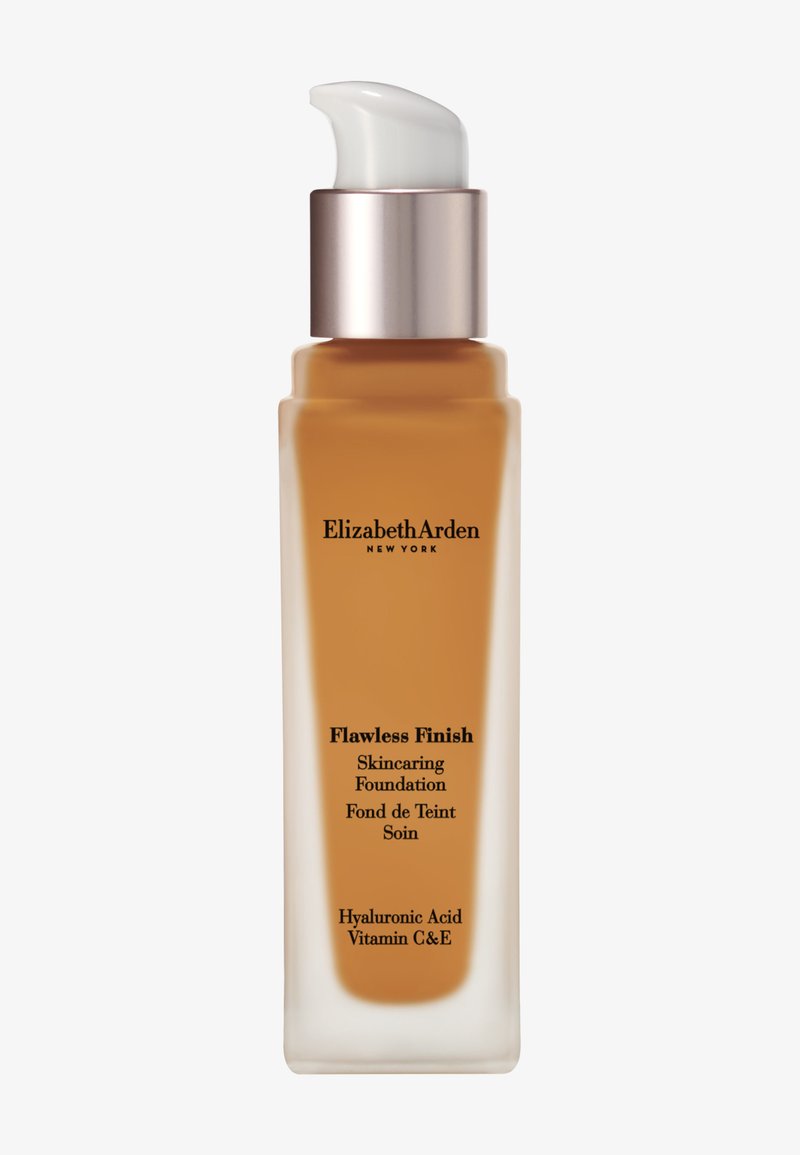 Fond de teint Flawless Finish dans un flacon en verre transparent. Distributeur à pompe, liquide beige, texte noir, capuchon argenté, mentionne de l'acide hyaluronique et des vitamines C et E.