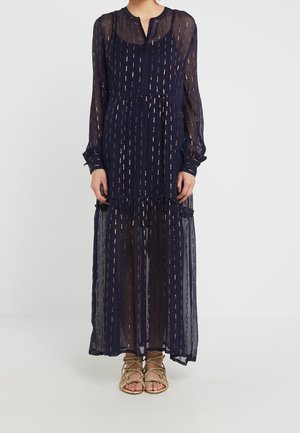 Vestido maxi de manga larga azul marino con tela transparente y rayas metálicas verticales, combinado con sandalias trenzadas doradas en una persona de pie.