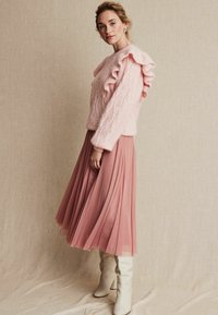 Różowy sweter z dzianiny z falbanami na ramionach; w połączeniu z zwiewną, plisowaną spódnicą w odcieniu różowego i białymi botkami za kolano. Miękkie, teksturowane materiały.