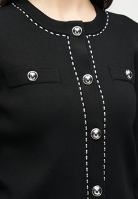 Μαύρο πλεκτό κάρντιγκαν με ασημένια στρογγυλά κουμπιά, λευκή ραφή κατά μήκος της γραμμής των κουμπιών και του λαιμού, και δύο λεπτομέρειες στο στήθος με καπάκια και κουμπιά.