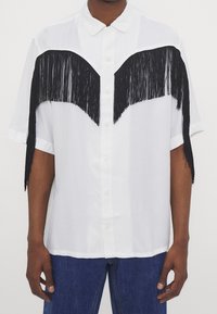 Camicia bianca con bottoni e dettagli in frange nere sul petto e sulle maniche. Realizzata in tessuto dalla texture liscia.