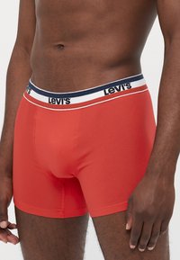 Raudoni medvilniniai bokseriai su tamsiai mėlyna ir balta elastinga juosta, ant kurios yra "Levi's" logotipas. Lygus paviršius, prigludęs dizainas, iki vidurio šlaunies ilgio.