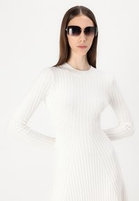 Robe tricotée blanche à côtes avec des manches longues et un col rond. Présente des motifs texturés diagonaux. De grandes lunettes de soleil oversize complètent le look.