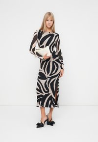 Robe à manches longues beige avec un motif zèbre noir, présentant une silhouette ajustée et une fente sur le côté. Talons noirs avec nœud et une pochette crème.