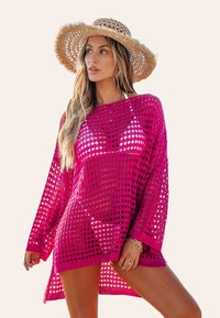 Różowa siateczkowa narzutka z dużymi perforacjami, szerokimi rękawami i luźnym fasonem, zestawiona ze słomkowym kapeluszem. Posiada swobodny design odpowiedni na plażę.