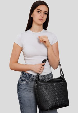 Junge Frau mit langen dunklen Haaren, trägt ein weißes T-Shirt und Jeans, hält eine große schwarze gesteppte Handtasche am Henkel.