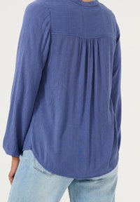 Blouse bleue à manches longues avec un tissu texturé, dos plissé et ourlet incurvé, associée à un jean bleu clair.