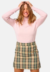 Femme souriante avec les mains dans les cheveux, portant un pull col roulé rose clair et une mini-jupe carrée beige, sur fond blanc.
