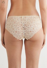 Beiga spetsunderkläder med blommönster, vågiga kanter och en semi-genomskinlig textur, visade från en bakre vinkel.