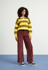 Geel en bruine geblokte gebreide trui met witte kraag, gecombineerd met maroon corduroy wijde broek en zwarte sneakers.