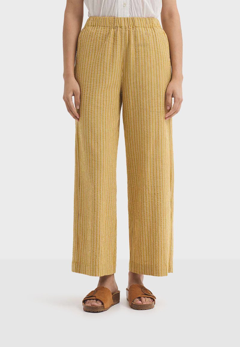 Femme portant un pantalon large rayé jaune et blanc, des sandales marron à enfiler et une chemise blanche boutonnée rentrée dans le pantalon.