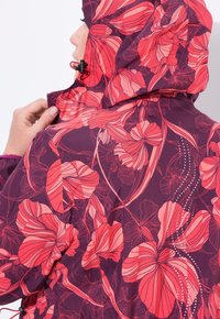 Žena upravující zip na bundě s kapucí s výrazným růžovým a červeným květinovým vzorem na tmavě fialovém pozadí.