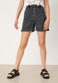 Mujer con pantalones cortos de mezclilla negros de cintura alta con bolsillos frontales, una blusa de encaje blanca y sandalias negras de tacón grueso y abierta en la parte delantera.