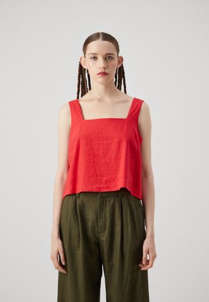 Rød kortermet tanktop med firkantet utringning, lavet af tekstureret stof. Parret med løse olivengrønne bukser. Modellen har fletninger.