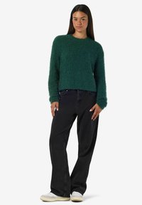 Zelen pulover z dolgimi rokavi in okroglim izrezom, v kombinaciji s črnimi ohlapnimi kavbojkami in belimi čevlji.