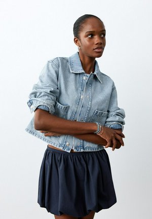 Mujer de piel oscura y cabello recogido que lleva una chaqueta de mezclilla clara con las mangas remangadas, una falda azul marino y pulseras de cuentas en la muñeca.