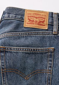 Blue denim jeans featuring a tan leather logo patch at the waistband, orange stitching, and subtle fading along the fabric.