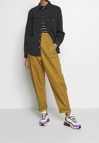 Svart denimskjorta med knapppanel, randig crop top, tan plisserade byxor och färgglada sneakers med lila och gula detaljer.