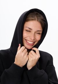 Zwarte hoodie van zacht materiaal, met een grote capuchon en geribbelde manchetten, met een close-up van een persoon die de capuchon vasthoudt.