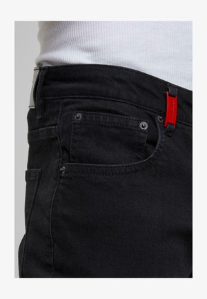 Lähedane vaade mustade denim pükste taskule ja vöökohale koos punase "2Y PREMIUM" sildiga, kantud valge ribakojaga pluusiga.