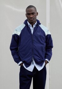 Navy windbreaker jacket with light blue accents, front zipper, and elastic cuffs. Worn over a white shirt, lending a layered look.