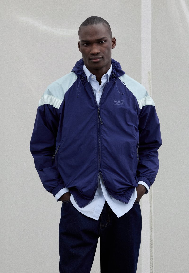 Navy windbreaker jacket with light blue accents, front zipper, and elastic cuffs. Worn over a white shirt, lending a layered look.