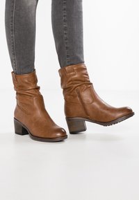 Bottines en cuir brun avec un design décontracté, fermeture éclair sur le côté, bout arrondi et un talon épais, associées à un jean skinny gris.