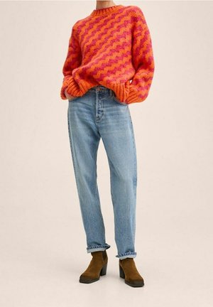 Personne portant un pull orange et rose à motifs en zigzag, un jean taille haute bleu clair retroussé aux chevilles, et des bottines marron à talons, debout les mains dans les poches.
